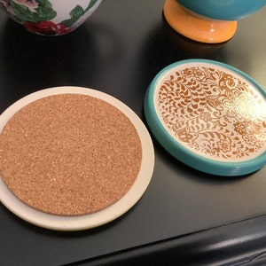 May include: Two round coasters, one with a cork top and a white ceramic base, and the other with a turquoise ceramic base and a white top with a gold floral design.