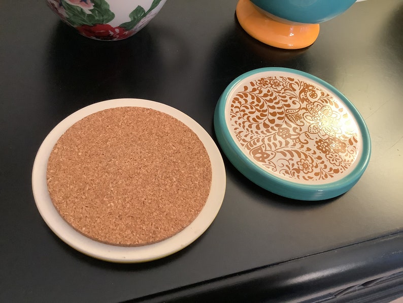 May include: Two round coasters, one with a cork top and a white ceramic base, and the other with a turquoise ceramic base and a white top with a gold floral design.