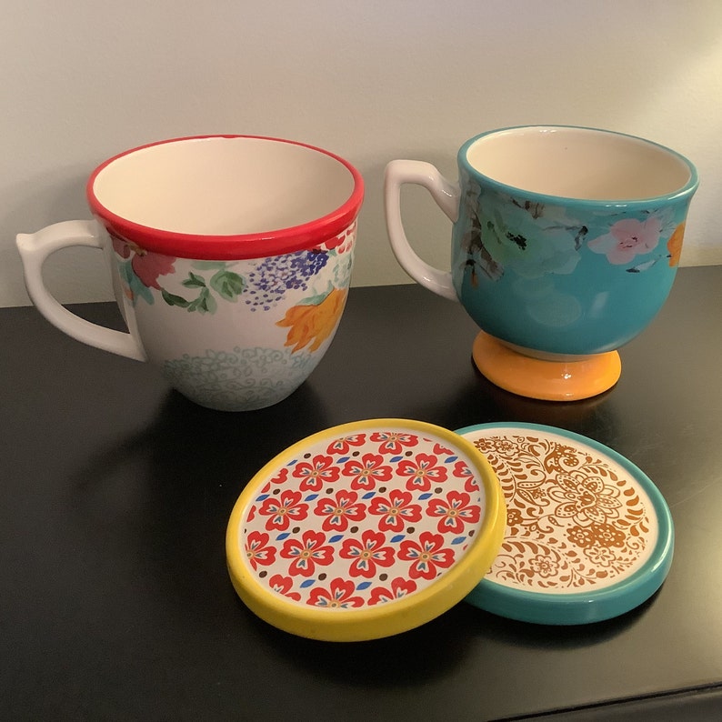 May include: Two floral patterned mugs with a red rim and a teal rim, sitting on top of two matching coasters with a yellow rim and a teal rim.