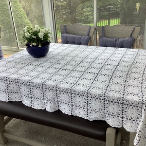 May include: White crocheted tablecloth with a scalloped edge, covering a wooden table with four chairs. The chairs have blue cushions and the table has a blue ceramic vase with white flowers.