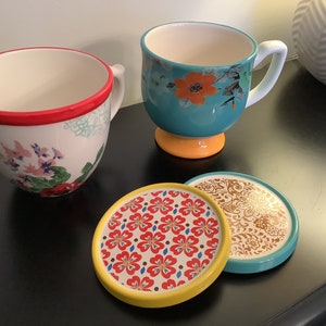 May include: Two floral patterned mugs with handles and a yellow base, and two matching coasters with floral patterns in red, yellow, and blue.