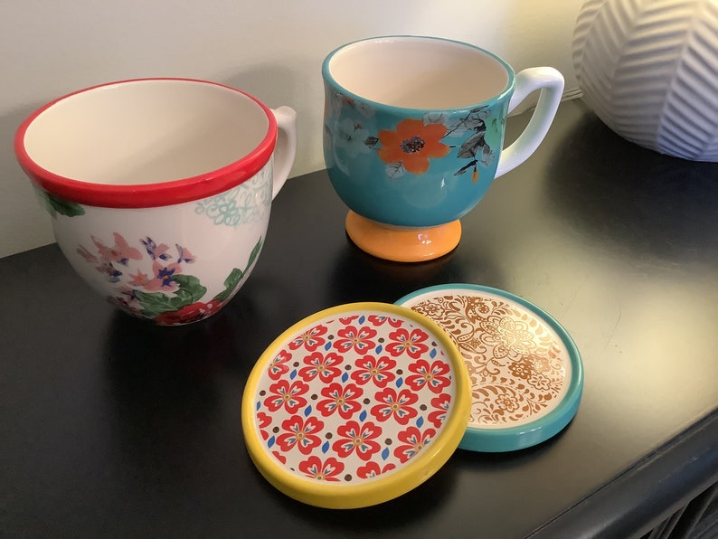 May include: Two floral patterned mugs with handles and a yellow base, and two matching coasters with floral patterns in red, yellow, and blue.