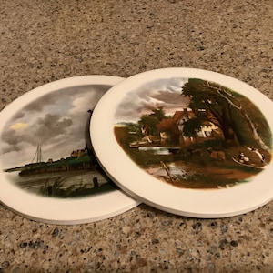 May include: Two round ceramic coasters with painted images of landscapes. One coaster features a river scene with a house and trees, while the other shows a cityscape with a bridge and boats.