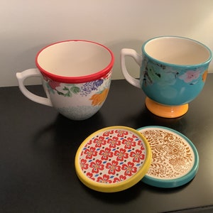 May include: Two ceramic mugs with floral patterns and a red rim. One mug is teal with a yellow base and the other is white with a yellow base. Two coasters with floral patterns are in the foreground.