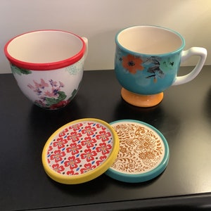 May include: Two floral patterned teacups with matching coasters. One cup is turquoise with a yellow base and the other is white with a red rim. The coasters are round and feature a variety of floral patterns.