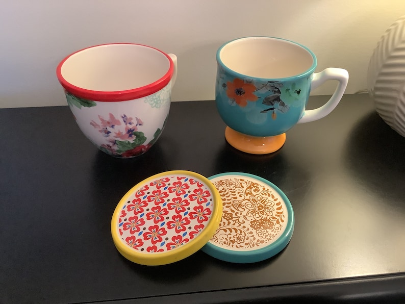 May include: Two floral patterned teacups with matching coasters. One cup is turquoise with a yellow base and the other is white with a red rim. The coasters are round and feature a variety of floral patterns.