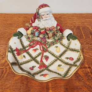 May include: A ceramic Santa Claus-shaped serving dish with a white and gold rim. The dish features a detailed design of fruits, berries, and a red ribbon. Santa's face and hat are red and white, with green gloves.