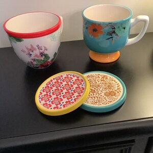 May include: Two floral patterned mugs with red and yellow accents, sitting on two colorful coasters with floral patterns. The coasters are yellow, turquoise, and brown.