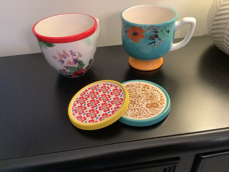 May include: Two floral patterned mugs with red and yellow accents, sitting on two colorful coasters with floral patterns. The coasters are yellow, turquoise, and brown.