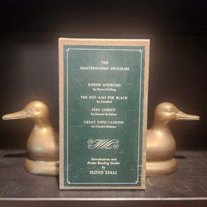 May include: A set of bookends featuring two gold-colored duck figurines flanking a green book with the title "The Masterworks Program." The book lists titles like "Joseph Andrews" and "Great Expectations."