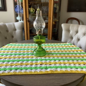 May include: A crocheted table runner with yellow, white, and green stripes. The runner is on a wooden table with a green glass oil lamp in the center.