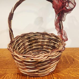 May include: A round, woven basket with a handle and a decorative burgundy ribbon. The basket is made of brown and tan woven material. The handle is wrapped with a darker brown material.