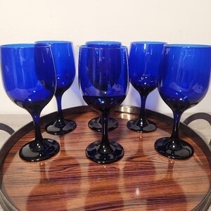 May include: A set of five cobalt blue glass wine goblets with rounded bowls and slender stems, arranged on a round serving tray featuring a dark brown wood grain pattern and a black metal rim with two side handles for easy carrying.