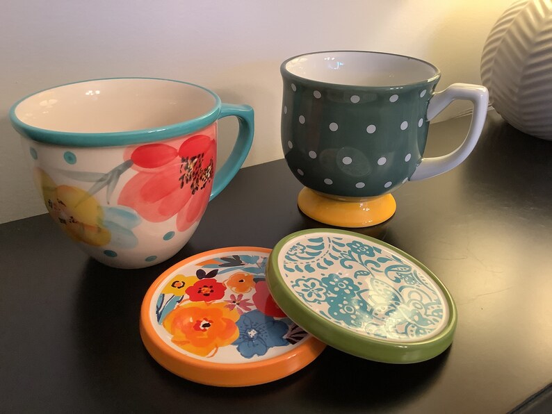 Pu&ograve; includere: Due tazze in ceramica con disegni floreali e a pois, e due sottobicchieri abbinati con motivi floreali. Le tazze sono sopra i sottobicchieri.
