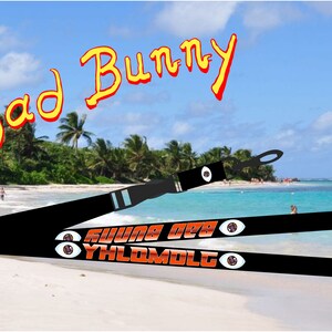 May include: Black lanyard with the text "YHLQMDLG" and "X100TO" in orange with a red eye graphic on each side. The lanyard is on a beach with a blue sky and palm trees in the background. The text "Bad Bunny" is written in yellow and red at the top of the image.