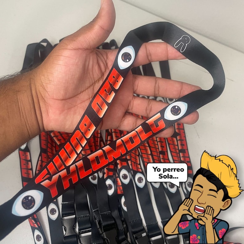 May include: A stack of black and white lanyards with red and white text that reads "YHLOMDLC" and "BAD OMEN". The lanyards have a black eye graphic with a red iris. One lanyard has a white bunny rabbit graphic. A person is holding one of the lanyards. A cartoon character with a straw hat is saying "Yo perreo Sola..."