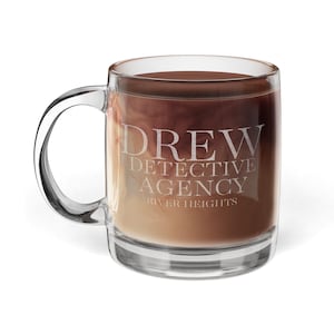 Puede incluir: Una taza de café de vidrio transparente con asa, llena de una bebida en capas. La taza tiene el texto "DREW DETECTIVE AGENCY RIVER HEIGHTS". La bebida tiene capas de tonos marrones y beige, creando un efecto degradado.