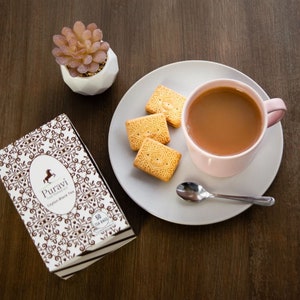 Könnte beinhalten: Eine Schachtel Puravi Ceylon Black Tea mit 50 Teebeuteln, eine rosa Teetasse mit Tee und drei Kekse auf einem weißen Teller.
