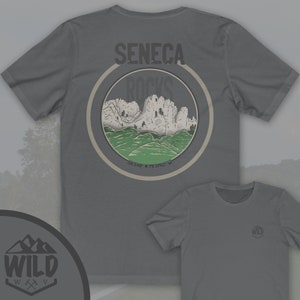 May include: Gray t-shirt with a circular graphic of a mountain range with the text "Seneca Rocks" and coordinates "38.348&deg; N 79.3762&deg; W". The graphic is surrounded by a brown circle. The t-shirt has a small logo on the bottom right corner that says "WILD" with a mountain range and an axe graphic.