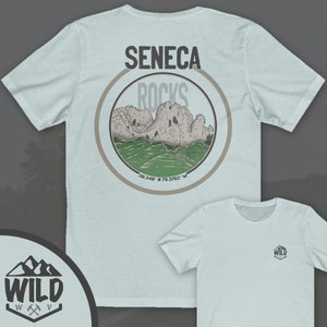 May include: A light blue t-shirt with a brown circle graphic on the back. The graphic features a mountain range with the text "Seneca Rocks" and coordinates "38.348&deg; N 79.3762&deg; W". The front of the shirt has a small logo with the text "WILD" in a mountain range design.