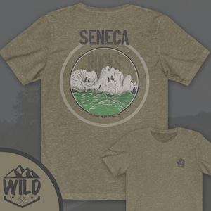 May include: A green and brown t-shirt with a graphic of Seneca Rocks, a mountain range in West Virginia. The graphic is in a circle with the text "Seneca Rocks" and coordinates "38.348 N 79.3762 W". The t-shirt has a small logo on the front that says "Wild" with a mountain range and an axe.