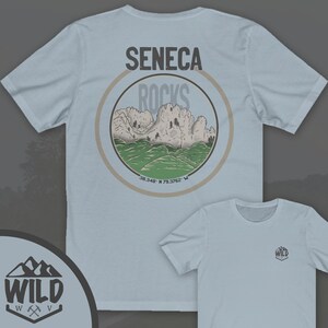 May include: Light blue t-shirt with a circular graphic on the back. The graphic features a mountain range with the text "Seneca Rocks" and coordinates "38.348 N 79.3762 W". The front of the shirt has a small logo with the text "WILD" and a mountain range graphic.