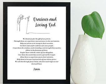 May include: A black framed print with a white background and a simple line drawing of a person praying. The text reads "Gracious and Loving God" and includes a prayer. The print is sitting on a white surface with a green plant in a white vase.