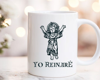 Könnte beinhalten: Weiße Keramiktasse mit einer Schwarzweißillustration eines Kindes mit erhobenen Armen und einem Heiligenschein. Der Text "YO REINARÉ" ist unter der Illustration gedruckt.