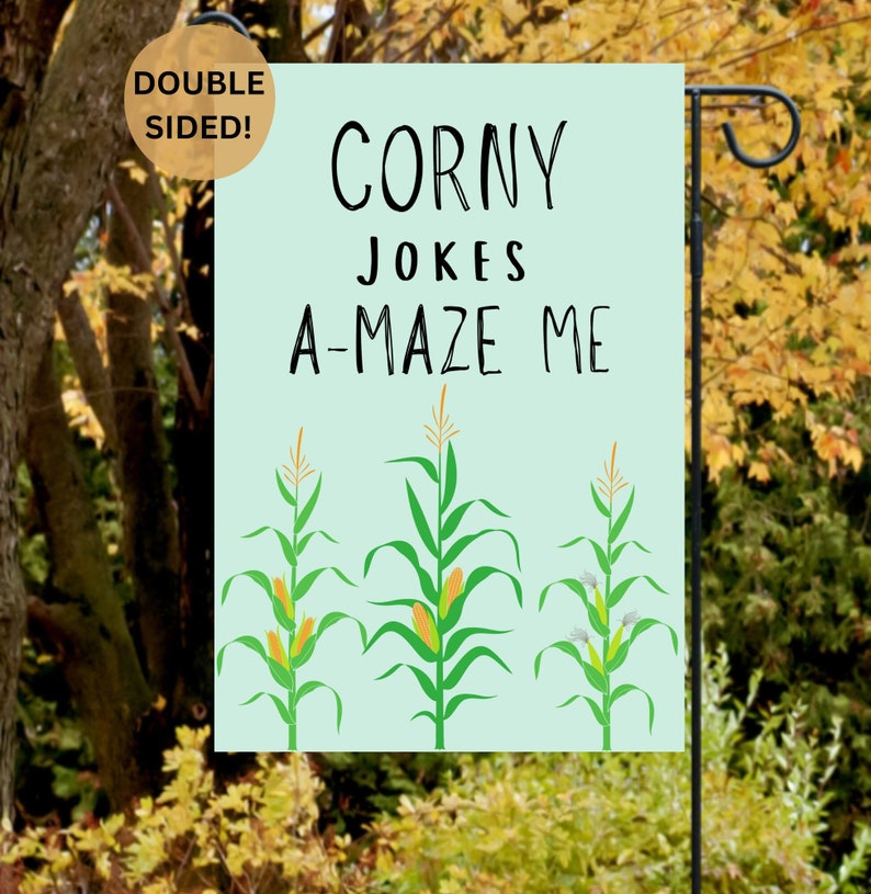 May include: A light blue garden flag with the text "Corny Jokes A-maze Me" and three cartoon corn stalks with ears of corn. The flag is double-sided.