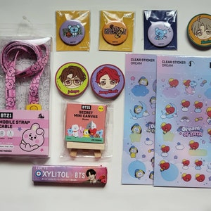May include: A collection of BT21 merchandise including a pink mobile strap cable, five circular character buttons, a secret mini canvas on a wooden easel, two sheets of clear dream stickers, and a Lotte Xylitol BTS gum pack with a portrait.