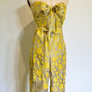 May include: A strapless yellow jumpsuit with a floral pattern and a bow detail at the bust. The jumpsuit has a matching belt and a fringed accent on one leg. The fabric has a metallic sheen.