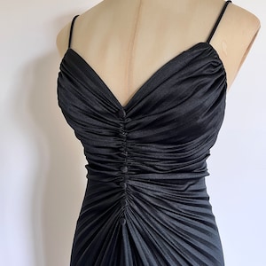 Op de afbeelding: Zwarte jurk met dunne bandjes, gepresenteerd op een mannequin. De jurk heeft een gerimpeld lijfje met een V-hals en een rij knopen. De rok is geplooid, wat een vloeiend effect creëert. Vintage stijl.