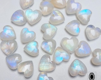 Pode incluir: Uma coleção de pedras preciosas em forma de coração de pedra da lua. As pedras são brancas com flashes iridescentes azuis e amarelos. As pedras preciosas estão espalhadas sobre uma superfície branca.