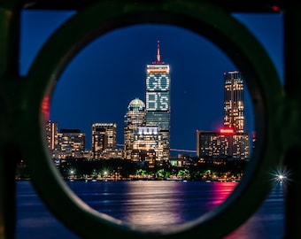 May include: A nighttime view of the Boston skyline through a circular opening in a metal fence. The tallest building in the center has the words "GO  B'S" illuminated on its side.