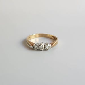 May include: A gold ring featuring three clear gemstones. The ring has a simple band and a classic design. The gemstones are round and likely diamonds. The ring is a piece of jewellery.