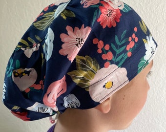 May include: A navy blue floral scrub cap with a pattern of pink, white, and peach flowers and green leaves. The cap is made of fabric and is designed to cover the head. The cap is worn by a person.