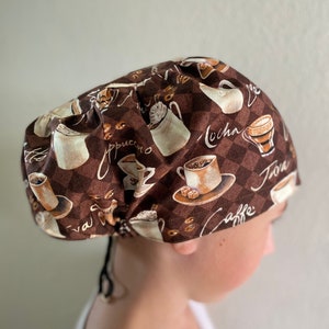 May include: A brown and white patterned scrub cap with coffee-themed illustrations. The cap has a checkered background with images of coffee cups, beans, and the words "caffe", "mocha", and "cappuccino". The cap is tied in the back with black ties.