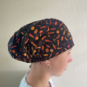 May include: A black scrub cap with a colorful print of various grilling foods, including hot dogs, hamburgers, and skewers. The cap is made of a soft, comfortable fabric and is perfect for keeping hair out of the way while cooking.