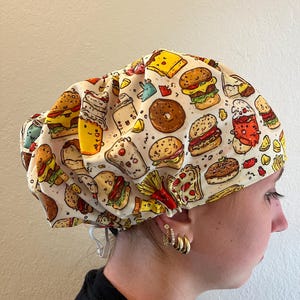 May include: A scrub cap featuring a cream-colored background with a repeating pattern of colorful fast food illustrations, including hamburgers, donuts, french fries, and various beverages. The cap has an adjustable elastic cord at the back.