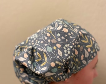 May include: A dark grey floral patterned bouffant scrub cap. The cap features a repeating pattern of leaves, flowers, and berries in shades of white, yellow, and light blue. The cap is gathered at the back and worn on the head.