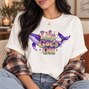 Pode incluir: Uma t-shirt de manga curta cor de creme com um gráfico de uma baleia com padrão de galáxia em tons de roxo e azul. O texto "Life Goes On" está impresso numa fonte degradê multicolorida no centro, rodeado por pequenas estrelas e uma lua crescente.