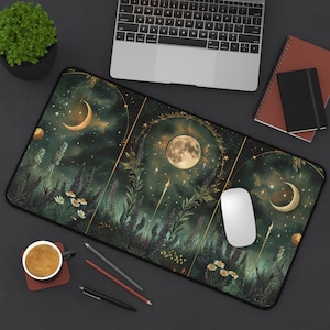 May include: A rectangular desk mat with a celestial design. It features a full moon, crescent moons, stars, and floral elements in green, gold, and white. A laptop, mouse, and stationery are on the dark surface.