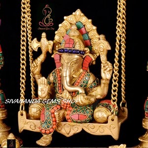 May include: A gold-colored statue of Ganesha, the Hindu god of wisdom and prosperity, sitting on a swing. The statue is decorated with colorful gemstones and intricate details. The swing is made of metal and has two chains.