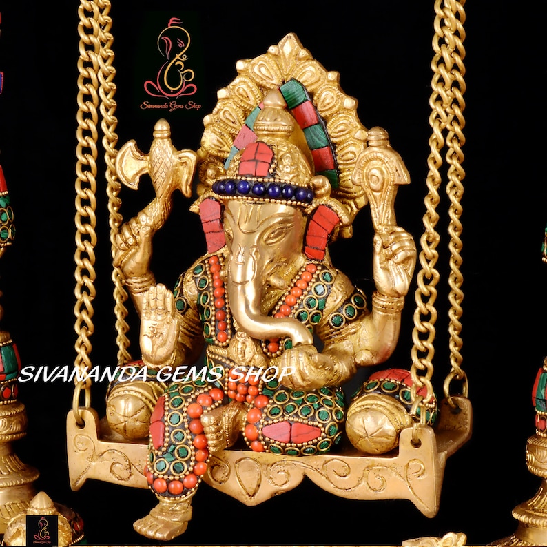 May include: A gold-colored statue of Ganesha, the Hindu god of wisdom and prosperity, sitting on a swing. The statue is decorated with colorful gemstones and intricate details. The swing is made of metal and has two chains.