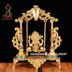 May include: A gold-colored metal statue of Ganesha, the Hindu god of wisdom and prosperity, sitting on a swing. The statue is decorated with colorful gemstones and is set on a base with intricate carvings. The swing is suspended from a decorative archway.