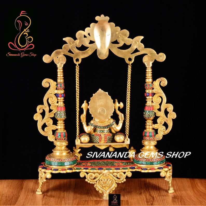 May include: A gold-colored metal statue of Ganesha, the Hindu god of wisdom and prosperity, sitting on a swing. The statue is decorated with colorful gemstones and is set on a base with intricate carvings. The swing is suspended from a decorative archway.
