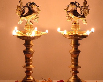 May include: Two gold metal oil lamps with bird finials and lit wicks. The lamps are on a surface with orange and red flower petals.