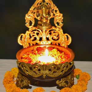以下が含まれることがあります： 神様の像、ろうそく、花びらが入った装飾的な真鍮製のランプ。オレンジ色のマリーゴールドの花のガーランドに囲まれています。白い表面に置かれています。