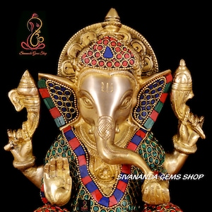 May include: A golden brass statue of the Hindu deity Ganesha, adorned with colorful inlaid stones. The statue features intricate details, including a large elephant head, multiple arms, and a decorative crown. The text "Sivananda Gems Shop" is visible.