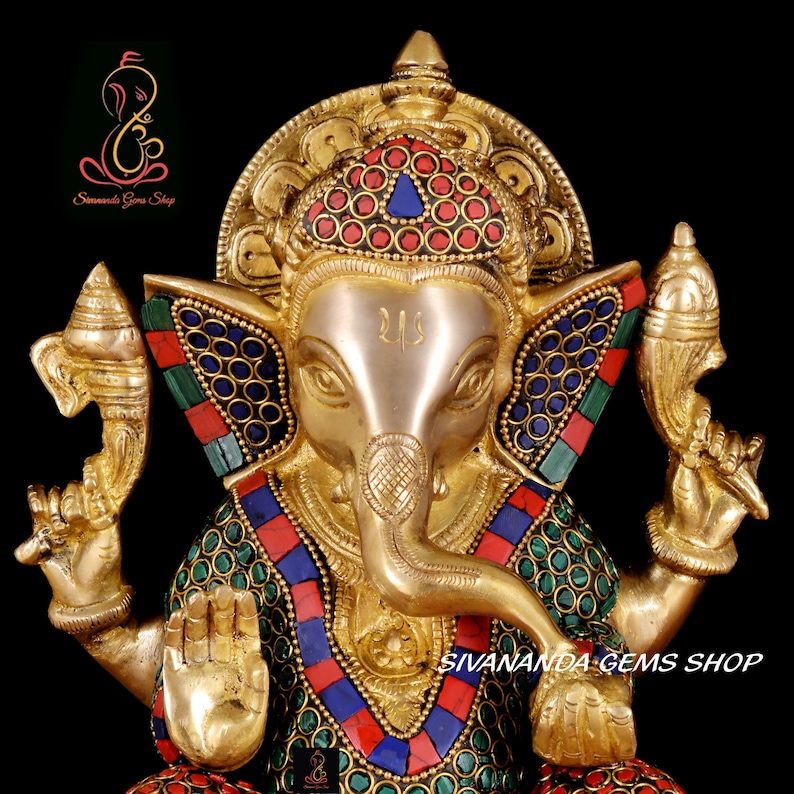 May include: A golden brass statue of the Hindu deity Ganesha, adorned with colorful inlaid stones. The statue features intricate details, including a large elephant head, multiple arms, and a decorative crown. The text "Sivananda Gems Shop" is visible.